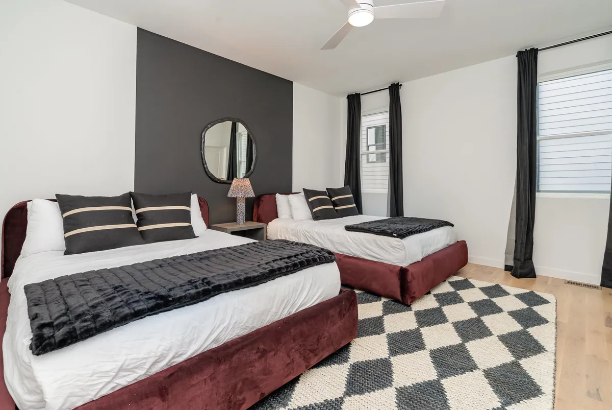 A cozy double bedroom with two beds featuring dark maroon upholstery, a geometric black and white rug, a decorative mirror, and soft light from a bedside lamp. The setting is warm, stylish, and perfect for friends or group stays.