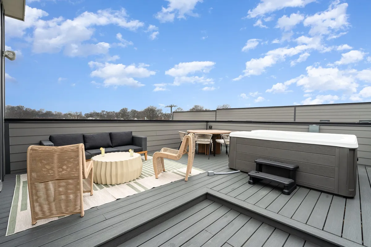 The image showcases a stylish rooftop patio at sunset, featuring a modern outdoor seating area, a bubbling hot tub, and a dining table set for gatherings, all with expansive city views. The ambiance is cozy and serene, inviting guests to relax, dine al fresco, and soak in the warm glow of the evening.