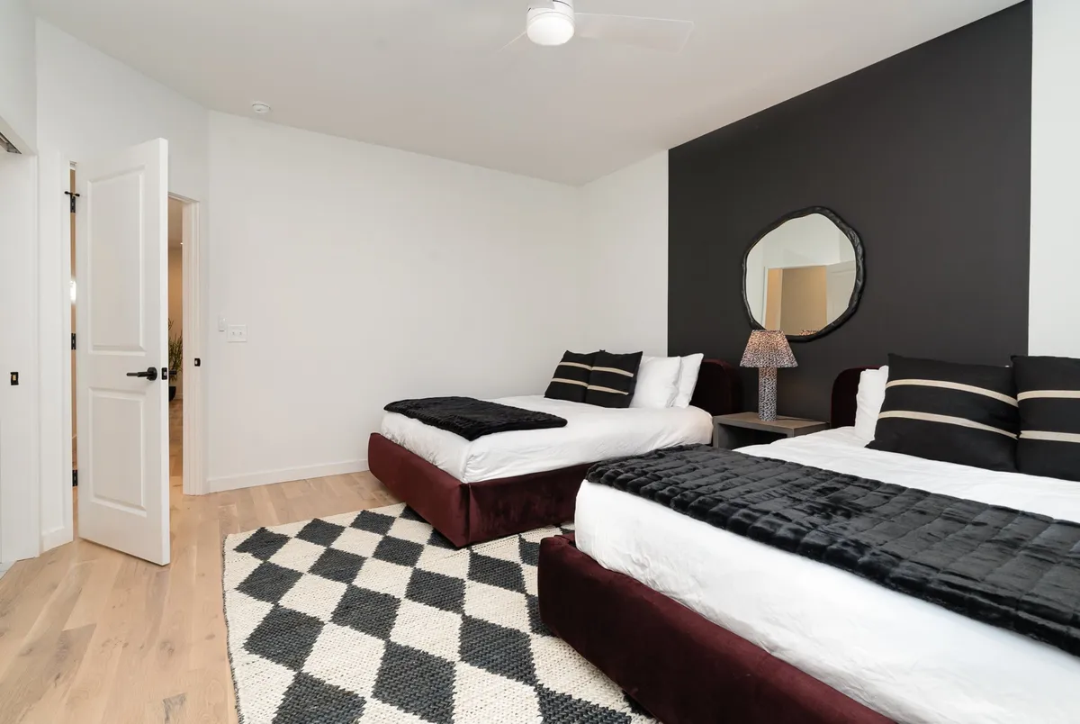 A modern guest room with two beds featuring dark burgundy bases, black and white striped pillows, and a large wall mirror. The geometric rug and open layout suggest ample accommodation for groups or families.