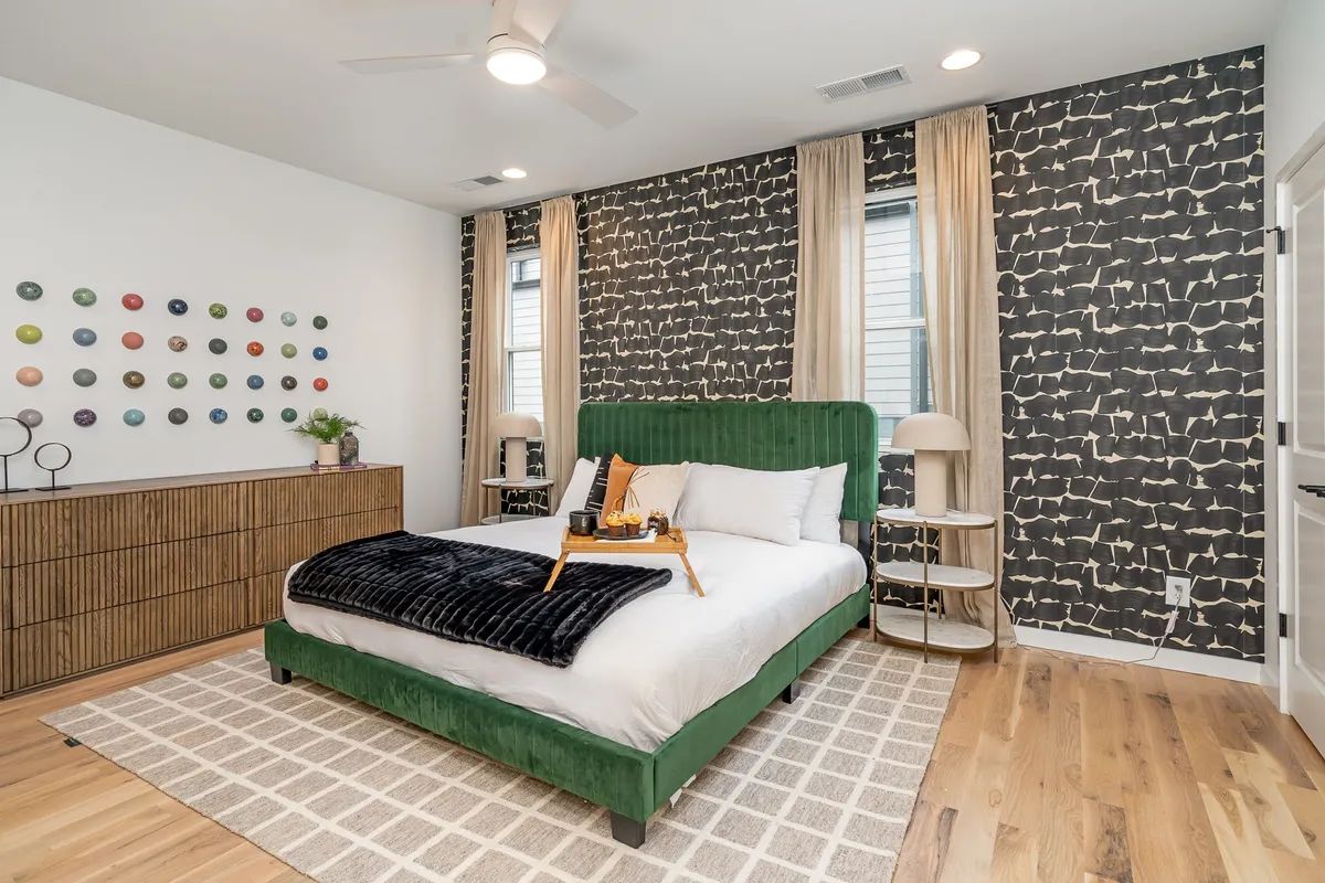 A stylish bedroom featuring a green upholstered bed, colorful wall art, a textured throw blanket, and natural wood furniture. Sunlight streams through the windows, creating an inviting and restful atmosphere.