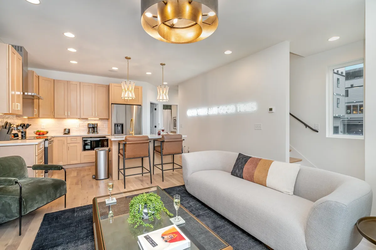 This image highlights a cozy and modern open-plan living space featuring a plush sofa, kitchen area with bar stools, and decorative lighting. Neon text adds a touch of personality and fun, while the layout effortlessly connects the kitchen and living area, reinforcing the theme of togetherness.